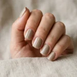 Photorealistic close-up of a hand with short square nails in warm sand greige with one fine white line running lengthways up each nail