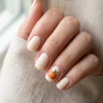 Photorealistic close-up of a hand with cream nails with one small round orange pumpkin and green stem on an accent nail