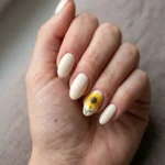 Photorealistic close-up of a hand with short almond nails painted soft cream, the ring fingernail featuring a single hand-painted yellow sunflower with a dark brown center and slightly uneven petals, soft diffused window light, clean neutral background, shot slightly from above, vertical 4:5 crop, sharp macro focus on the nails, realistic skin and cuticle detail, no text, no watermark