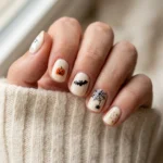 Mismatched short Halloween nails on cream base with ghost, pumpkin, bat, and web motifs